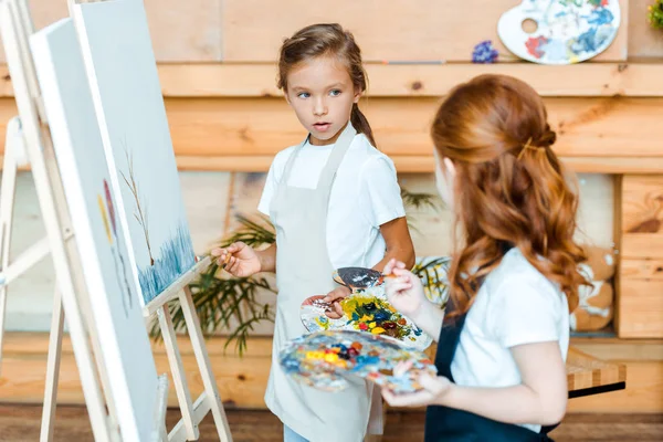 Selektiver Fokus des entzückenden Kindes, das das rothaarige Kind in der Nähe von Staffeleien in der Kunstschule betrachtet — Stockfoto