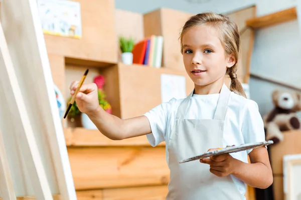 Selective focus of positive kid holding palette and paintbrush — Stock Photo