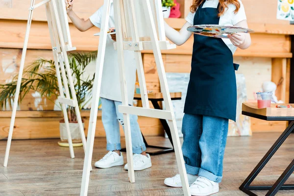 Cropped view of kids in aprons standing near easels — Stock Photo
