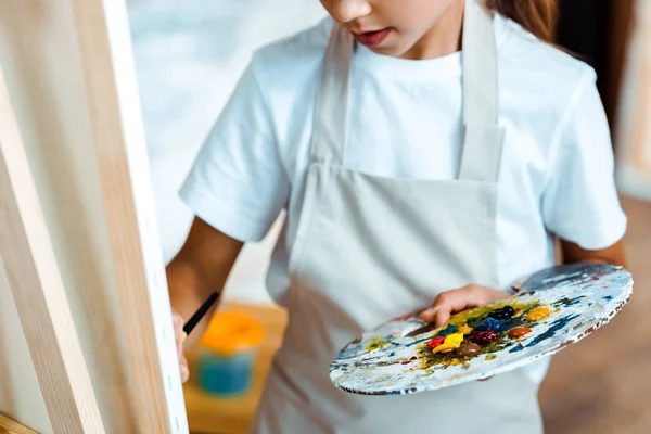 Ausgeschnittene Ansicht von Kind mit Palette, die neben Staffelei steht — Stockfoto