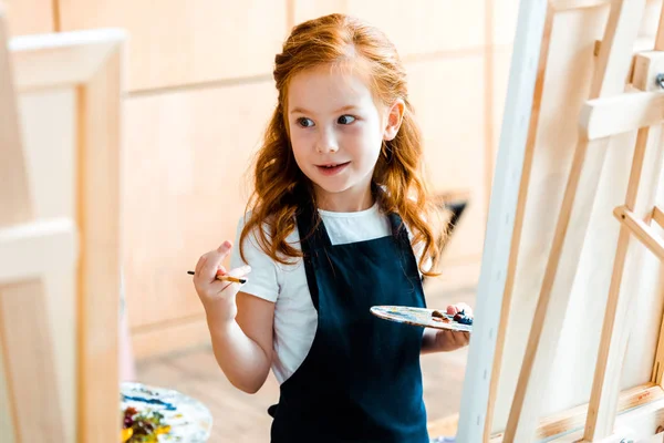 Messa a fuoco selettiva di cute rossa bambino tenendo tavolozza e guardando altrove — Foto stock