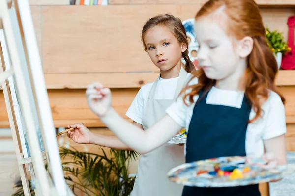 Foyer sélectif de mignon enfant regardant rousse enfant près du chevalet — Photo de stock