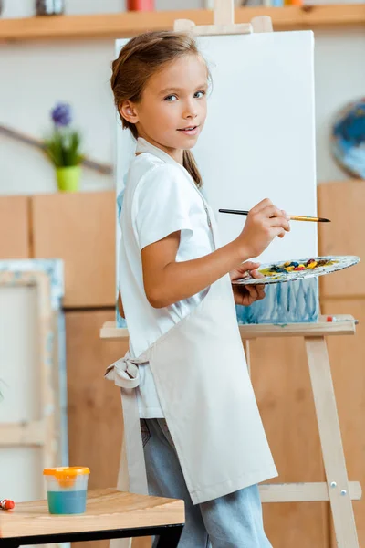 Cheerful kid in apron standing with palette and paintbrush near easel — Stock Photo