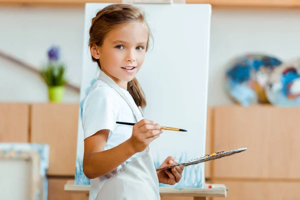 Happy kid in apron standing with palette and paintbrush near easel — Stock Photo