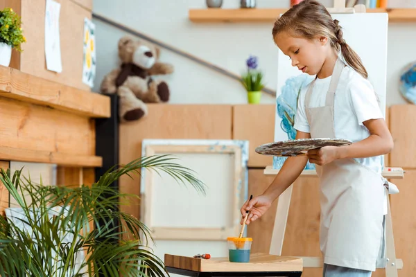 Mignon enfant dans tablier debout avec palette et pinceau près du chevalet — Photo de stock