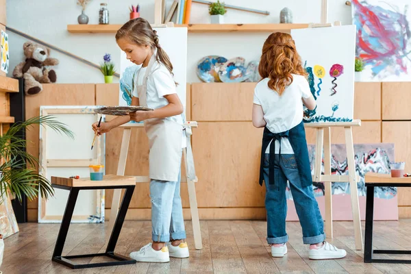 Back view of redhead kid holding palette while painting on canvas near child in art school — Stock Photo