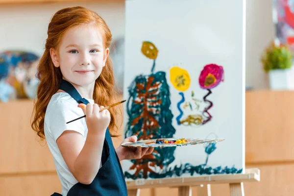 Souriant rousse enfant debout près de la peinture sur toile à l'école d'art — Photo de stock