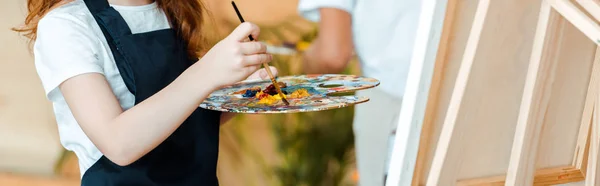 Panoramic shot of kid holding palette and paintbrush near easel — Stock Photo