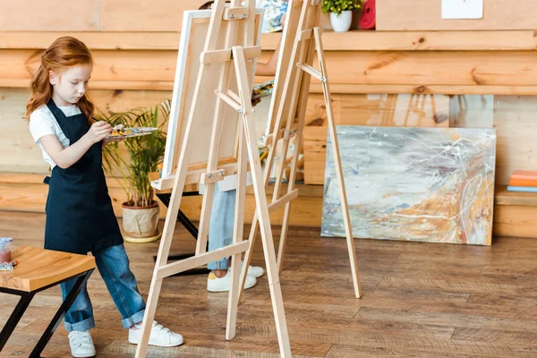 Милий рудий хлопець стоїть і малює в художній школі біля дитини — стокове фото
