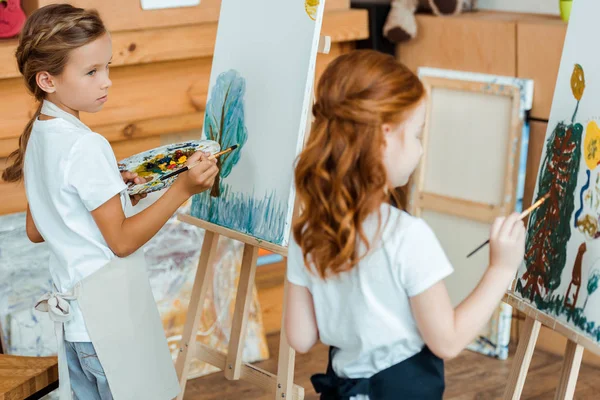 Selective focus of cute kid looking at child painting on canvas in art school — Stock Photo