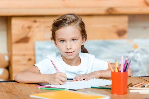 Selektiver Fokus des niedlichen Kindes, das in der Kunstschule auf Papier zeichnet und dabei in die Kamera schaut — Stockfoto