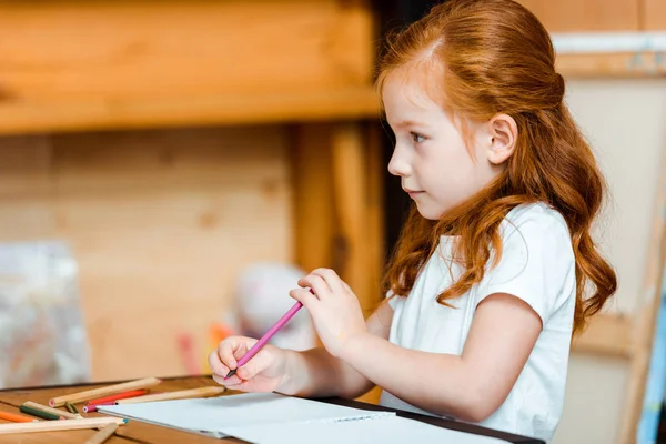 Lindo pelirrojo niño sosteniendo lápiz de color cerca de papel en la escuela de arte - foto de stock