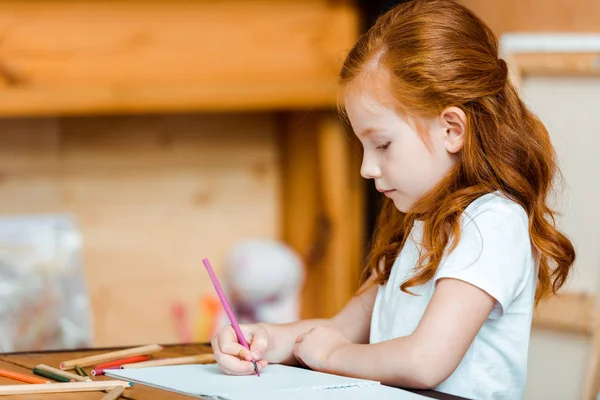 Lindo pelirroja niño celebración de lápiz de color mientras dibuja en papel - foto de stock