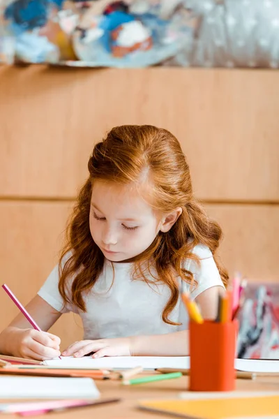 Enfoque selectivo de lindo niño pelirrojo sosteniendo lápiz de color mientras dibuja en papel - foto de stock