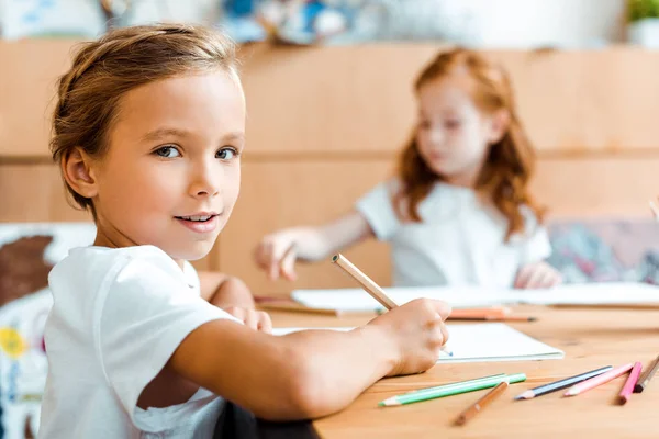 Foyer sélectif de l'enfant heureux avec crayon de couleur en regardant la caméra près de l'enfant — Photo de stock