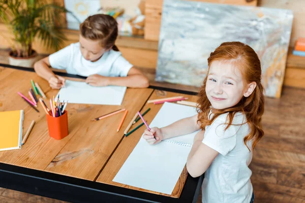 Selektiver Fokus des glücklichen rothaarigen Kindes, das in die Kamera schaut, während es in Kindernähe lächelt — Stockfoto