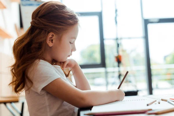 Side view of redhead child drawing on paper with color pencil — Stock Photo