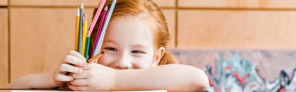 Plano panorámico de niña pelirroja sonriente sosteniendo lápices de color - foto de stock