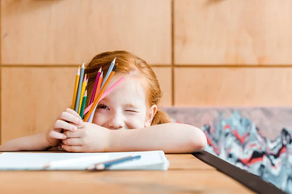 Селективное внимание милого рыжеволосого ребенка подмигивающего глазами, держа цветные карандаши — стоковое фото
