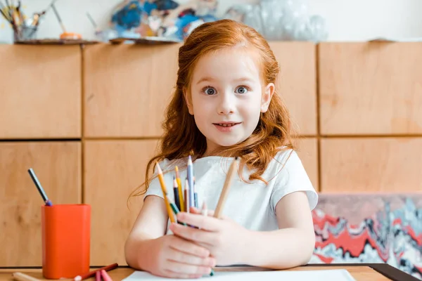 Niño pelirrojo excitado sosteniendo lápices de color en la escuela de arte — Stock Photo