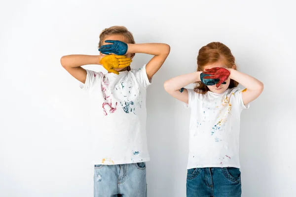 Lindos niños con pintura en las manos cubriendo caras mientras están de pie en blanco - foto de stock