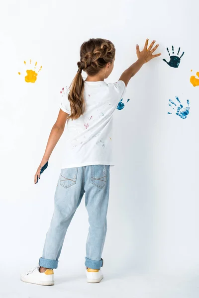 Vista trasera de niño en jeans poniendo la mano cerca de las huellas de la mano en blanco - foto de stock