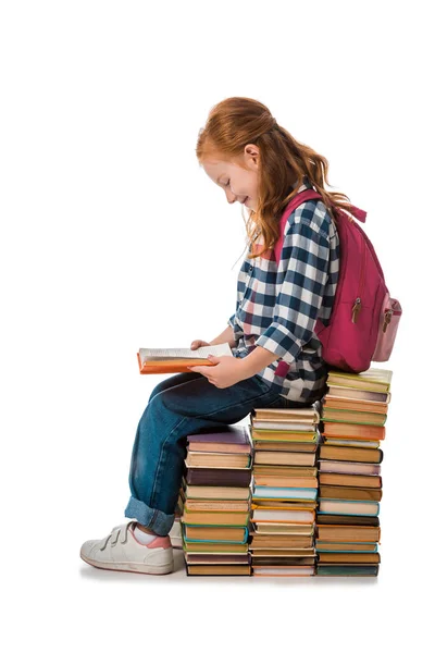 Cheerful redhead schoolkid smiling while sitting on books isolated on white — Stock Photo