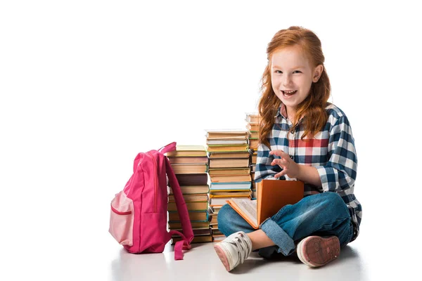 Alegre pelirroja escolar sentado cerca de libros y rosa mochila en blanco - foto de stock