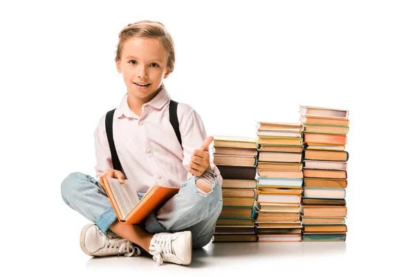 Fröhliches Kind, das mit überkreuzten Beinen in der Nähe von Büchern sitzt und Daumen hoch auf weiß zeigt — Stockfoto