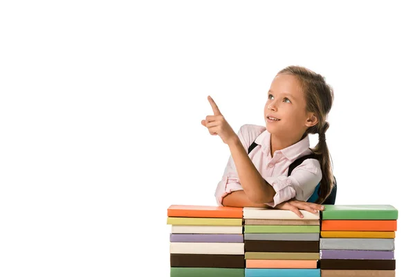 Positive schoolkid pointing with finger while looking away near colorful books isolated on white — Stock Photo