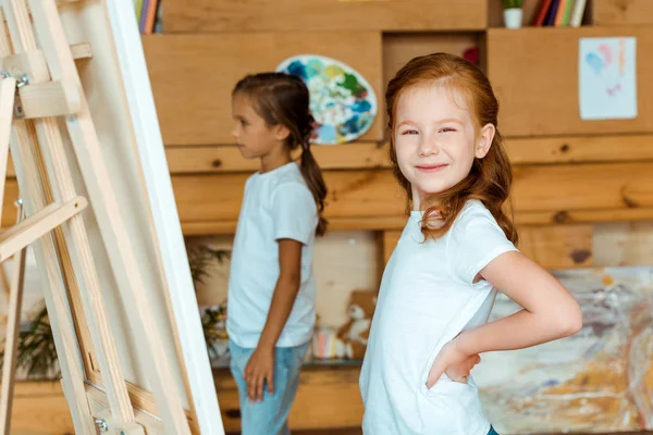 Enfoque selectivo de niño pelirrojo feliz de pie con la mano en la cadera cerca del niño en la escuela de arte - foto de stock