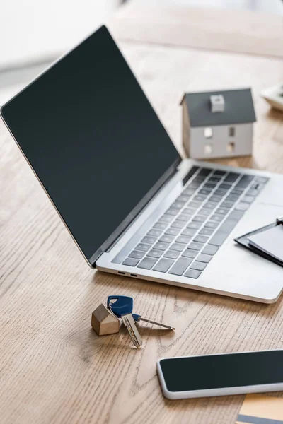 Laptop e smartphone con schermo bianco, chiavi e modello di casa su tavolo in legno — Foto stock