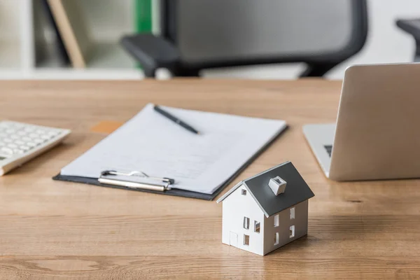 Clipboard and laptop near house model on wooden table in office — Stock Photo