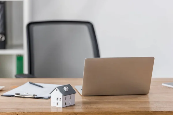 Modelo de casa, portátil y portapapeles con pluma en escritorio de madera cerca de la silla de oficina - foto de stock