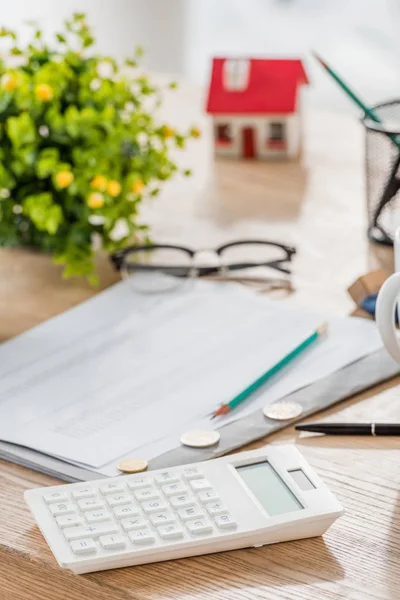 Enfoque selectivo de la calculadora cerca de monedas, papel, gafas, planta verde y modelo de casa en la mesa de madera - foto de stock