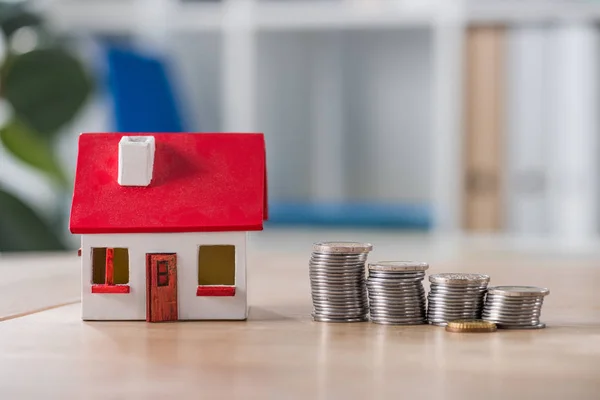 Modelo de casa cerca de pilas de monedas de oro y plata en la mesa de madera - foto de stock