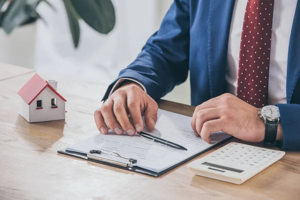 Vista parcial del hombre de negocios en el lugar de trabajo cerca de portapapeles, modelo de casa y calculadora - foto de stock