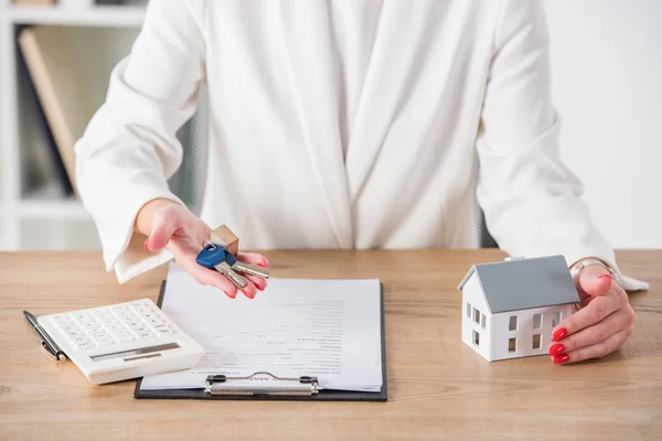 Partial view of businesswoman at workplace holding keys and touching house model near clipboard and calculator — Stock Photo