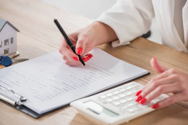 Zugeschnittene Ansicht einer Geschäftsfrau mit Taschenrechner und Schreiben im Vertrag in der Nähe von Hausmodell und Schlüsseln — Stockfoto