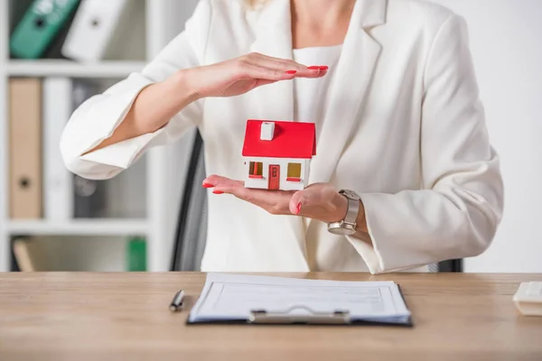 Partial view of businesswoman holding and covering house model with hand near clipboard — Stock Photo