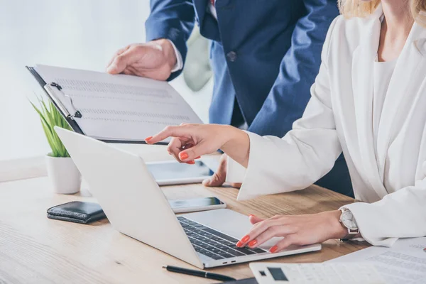 Cropped view of businessman holding clipboard near businesswoman using laptop at workplace — Stock Photo