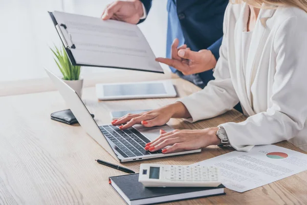 Vista parcial del hombre de negocios sujetando portapapeles cerca de la mujer de negocios utilizando el ordenador portátil en el lugar de trabajo - foto de stock