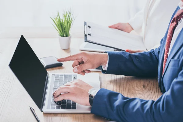 Cropped view of businessman pointing with finger at laptop monitor near colleague holding clipboard — Stock Photo