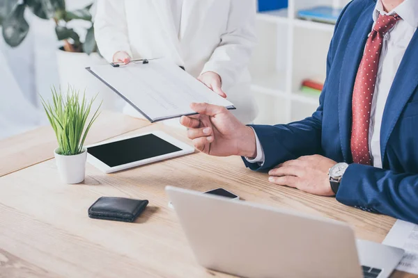 Vista recortada de la mujer de negocios sujetando portapapeles cerca de hombre de negocios sentado en el lugar de trabajo — Stock Photo