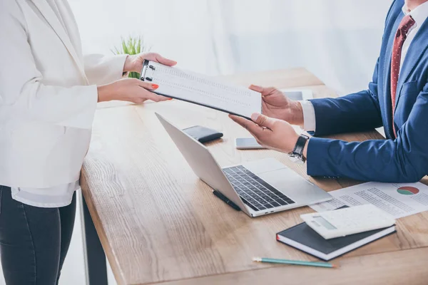 Cropped view of businesswoman giving clipboard to colleague sitting at workplace — Stock Photo