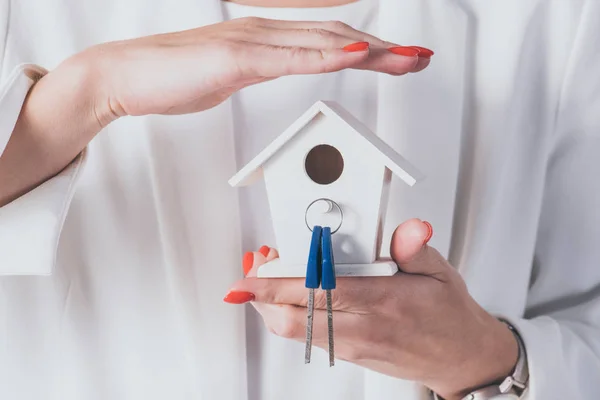 Vista cortada de empresária segurando e cobrindo modelo de casa e chaves com a mão — Fotografia de Stock