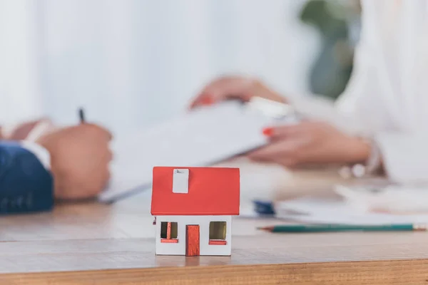 Selective focus of house model near businesswoman holding clipboard and client signing contract — Stock Photo