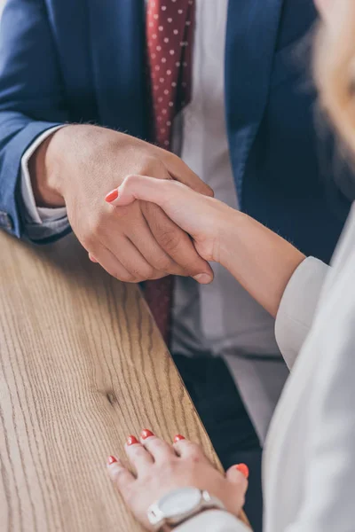 Vista recortada del hombre de negocios estrechando la mano con la mujer mientras está sentado en el escritorio de madera - foto de stock