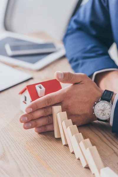 Cropped view of risk manager protecting house model from falling wooden blocks with hand — Stock Photo