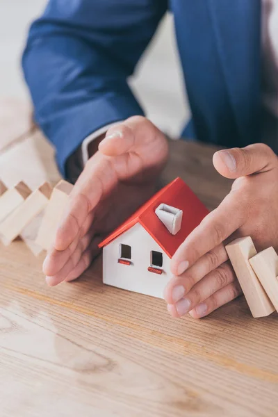 Cropped view of risk manager protecting house model from falling wooden blocks with hands — Stock Photo